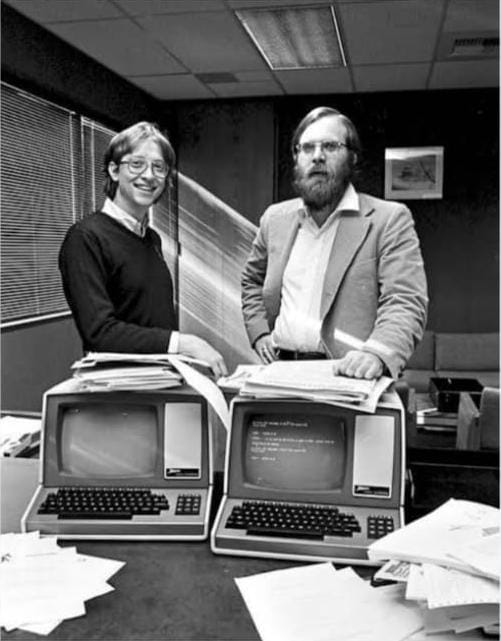 “Two men standing in an office with vintage computers on a desk, surrounded by papers, representing early days of personal computing.”